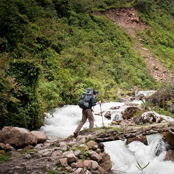 A man carrying hiking poles is walking over a bridge with water flowing under it. In front of him are mountains with green plants