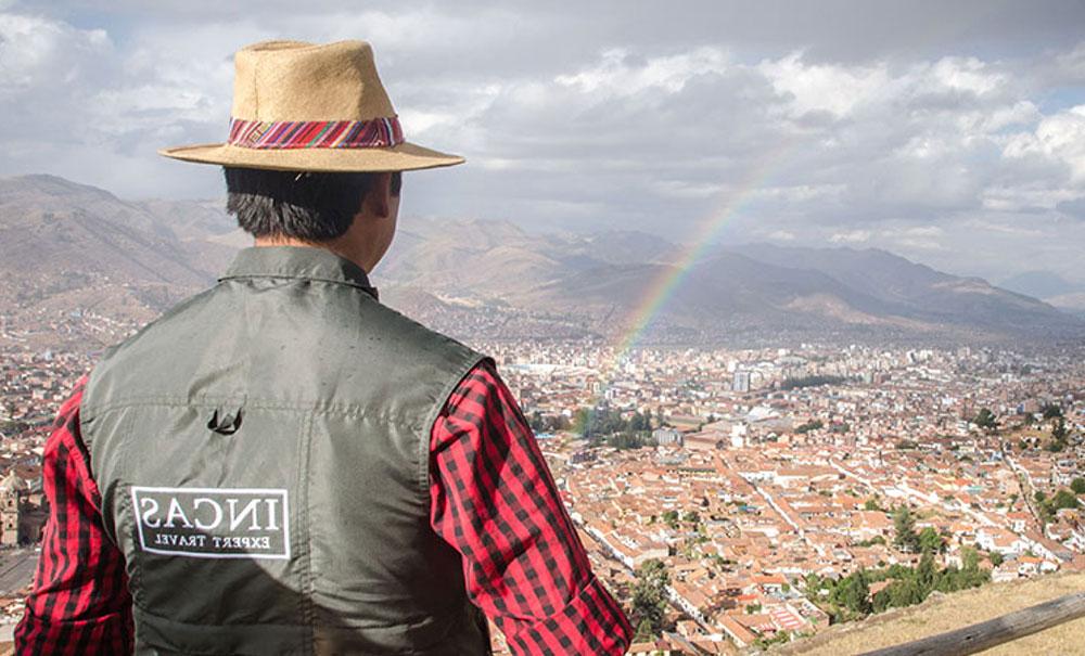 A man wearing an Inca Expert vest looking out at the whole city of Cusco. In the air, you can see a rainbow above the city