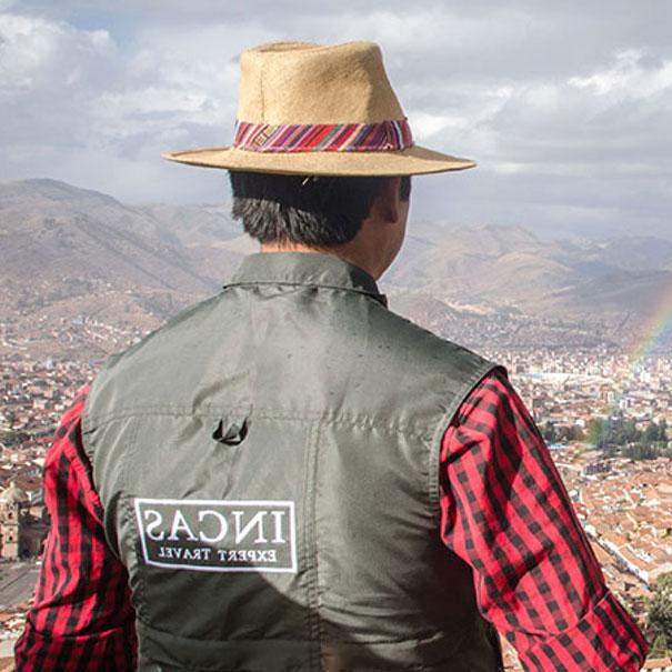A man wearing an Inca Expert vest looking out at the whole city of Cusco. In the air, you can see a rainbow above the city