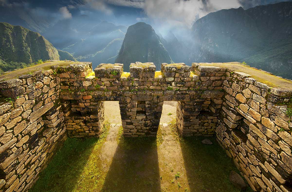 Afternoon sunlight shines through Inca windows at Machu Picchu with green mountains in the background.