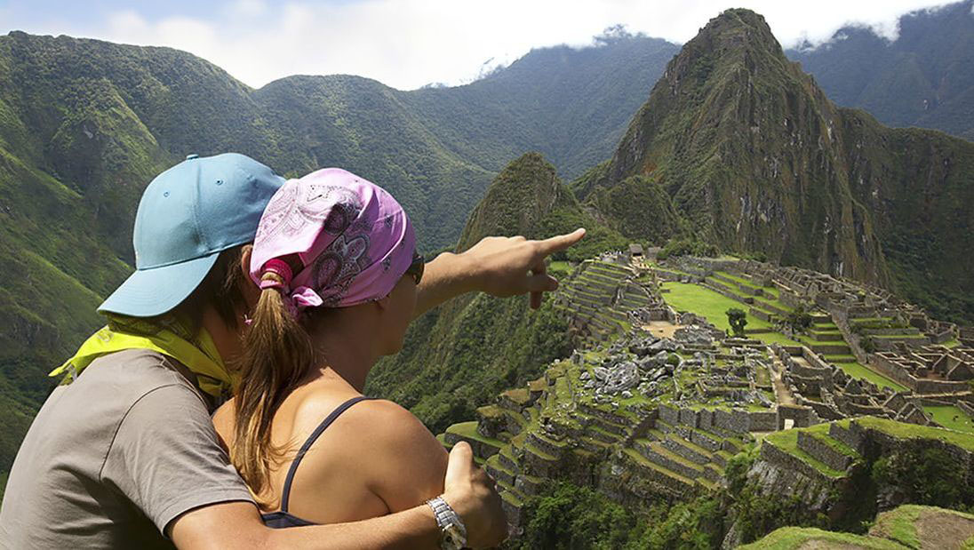 Couple point to the Machu Picchu ruins from lookout point with Huayna Picchu peak in the background.