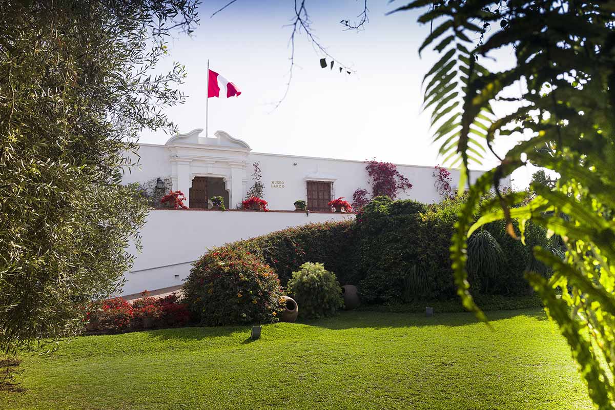 The white painted front facade of Museo Larco and the museum's lush gardens. A Peruvian flag is flown on top of the museum.