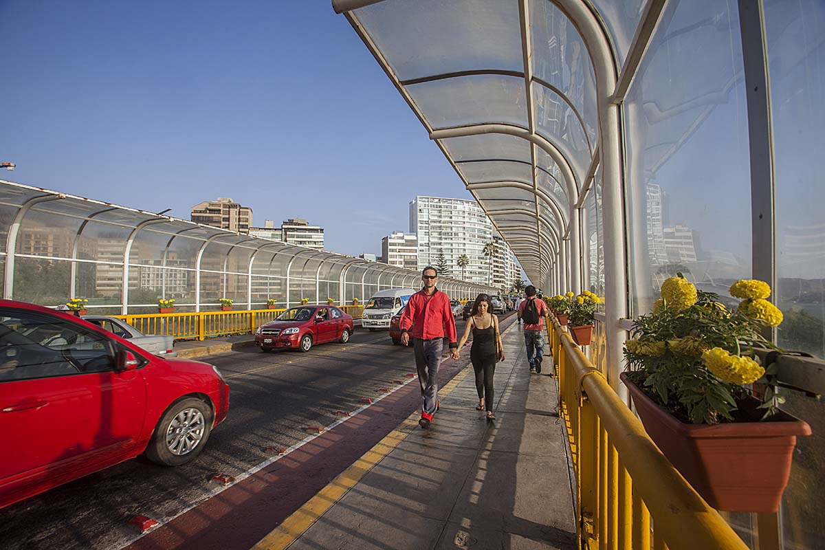 A man and woman in sports clothes walk hand in hand on the sidewalk of a bridge as cars pass.