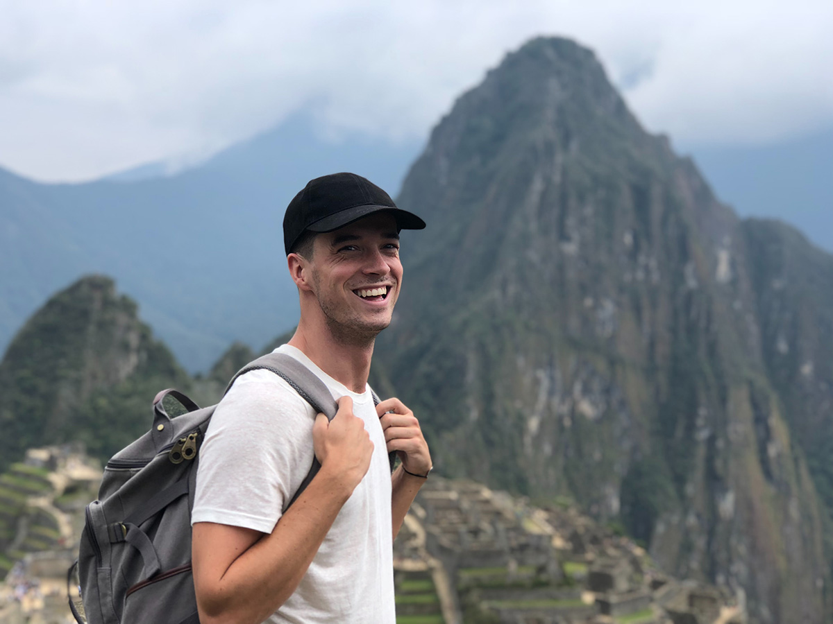 A man in a black baseball cap and white shirt with a gray backpack smiles in front of Machu Picchu.