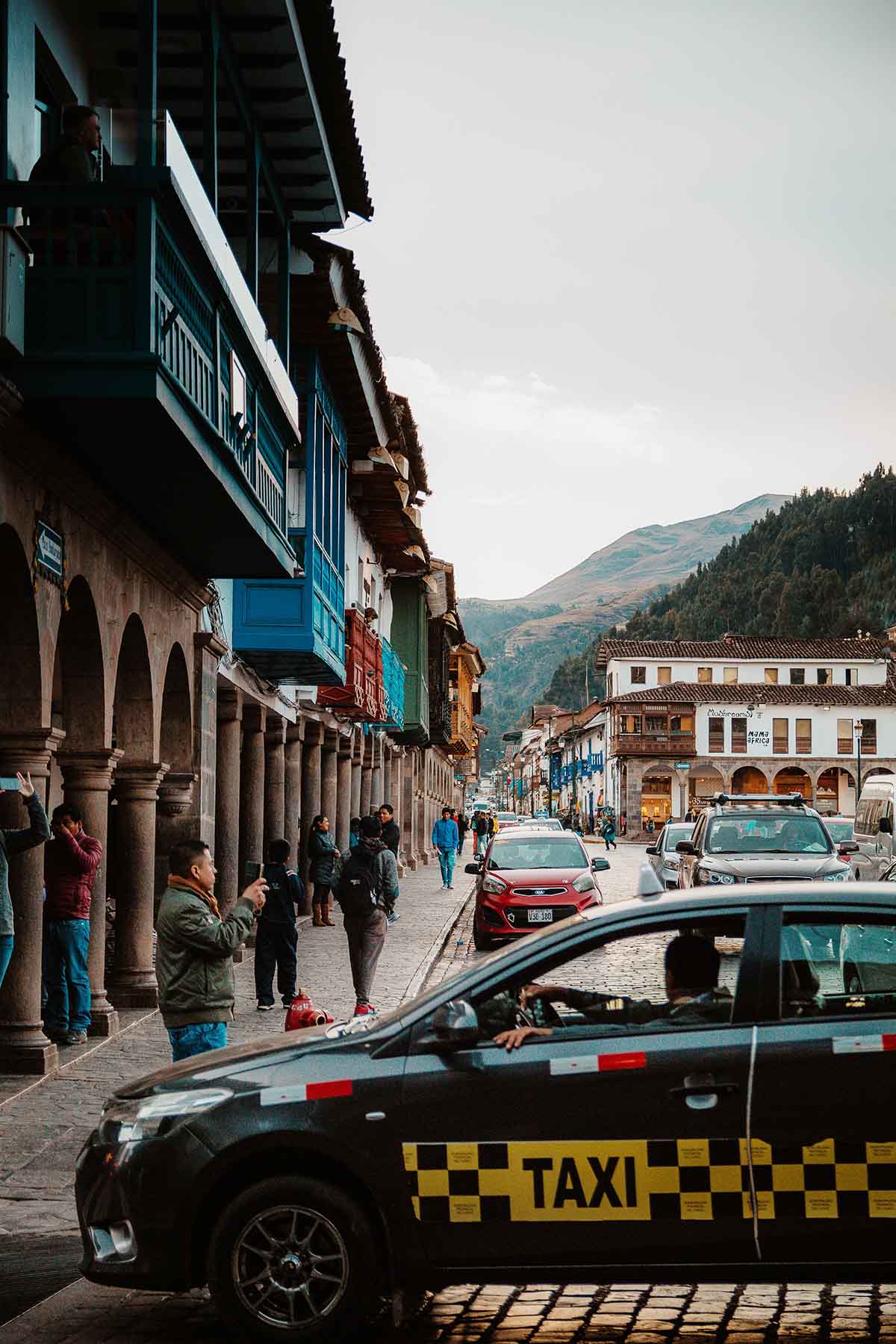 A taxi is parked in front of a row of arches on Cusco's Plaza de Armas as people walk by.