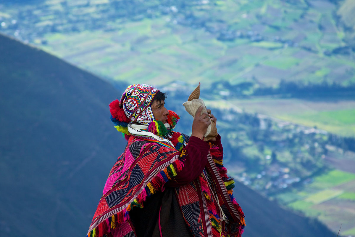 A shaman in a beaded hat with ear flaps and red poncho holds up a package in a rural landscape.
