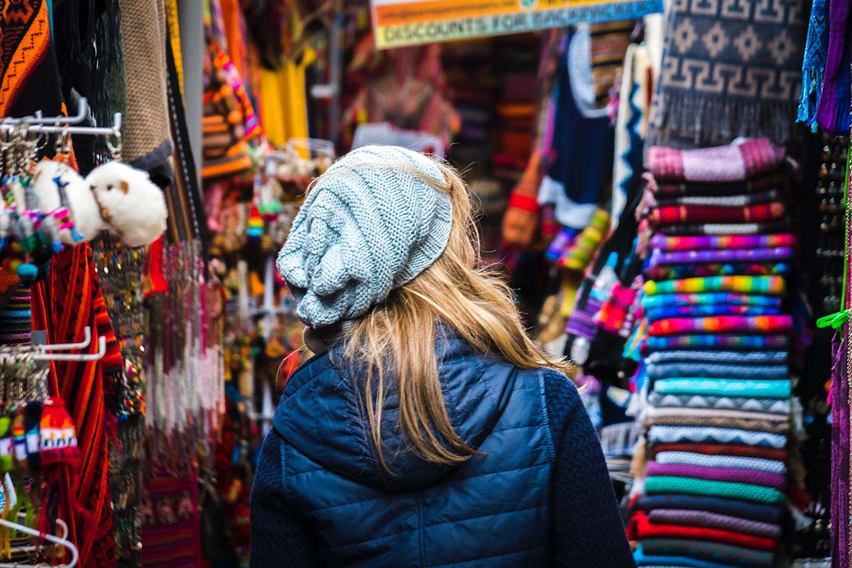 A young woman in a sagging gray knit beanie and light, puffy, dark blue jacket walks through a Peruvian souvenir market.