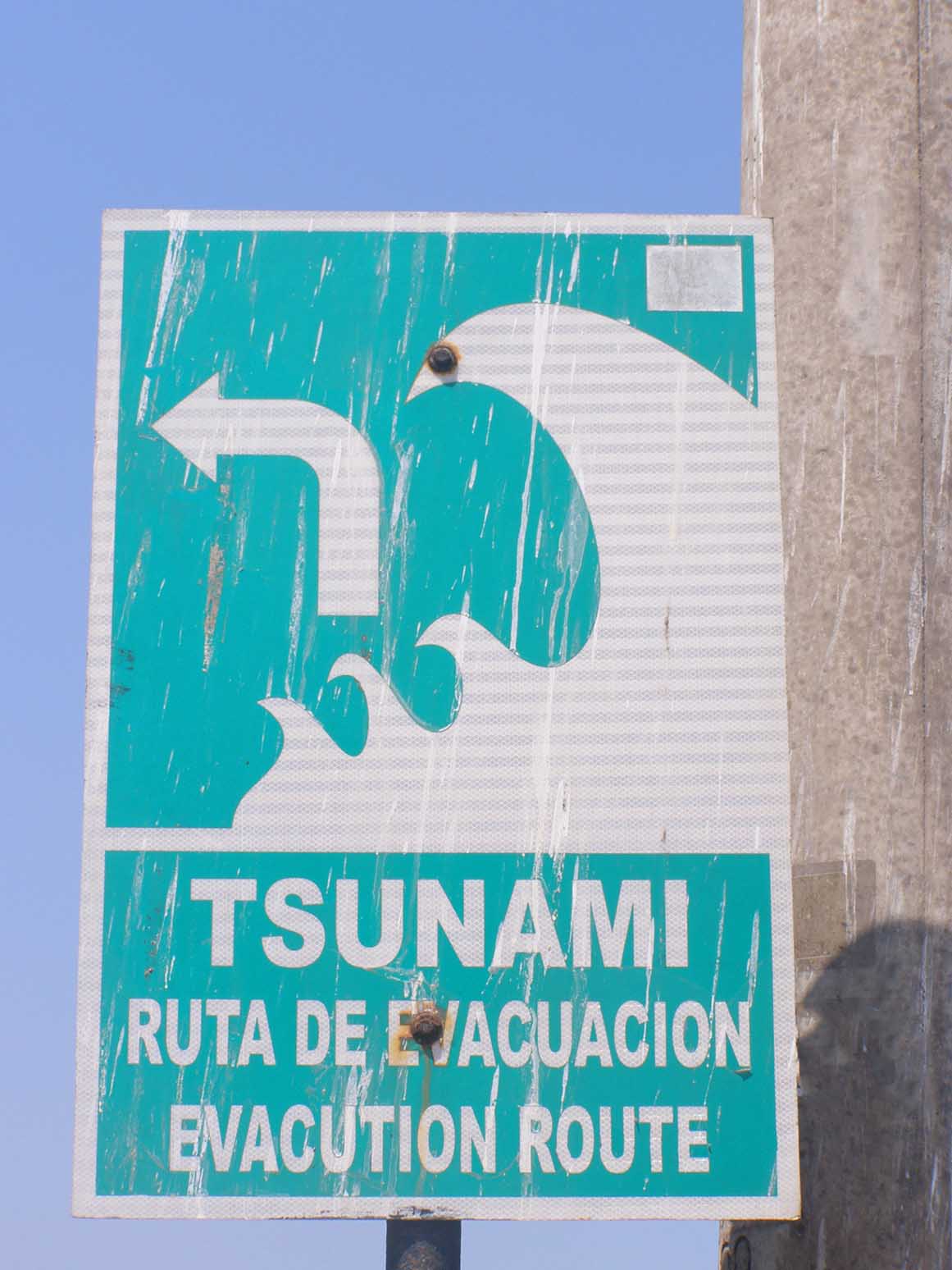 A green sign with a white wave and arrow says, "Tsunami, ruta de evacuación. Evacuation route."