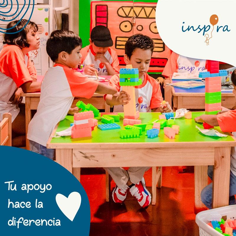 Three children sitting at a green table playing with large legos. There are two other tables behind with other children playing