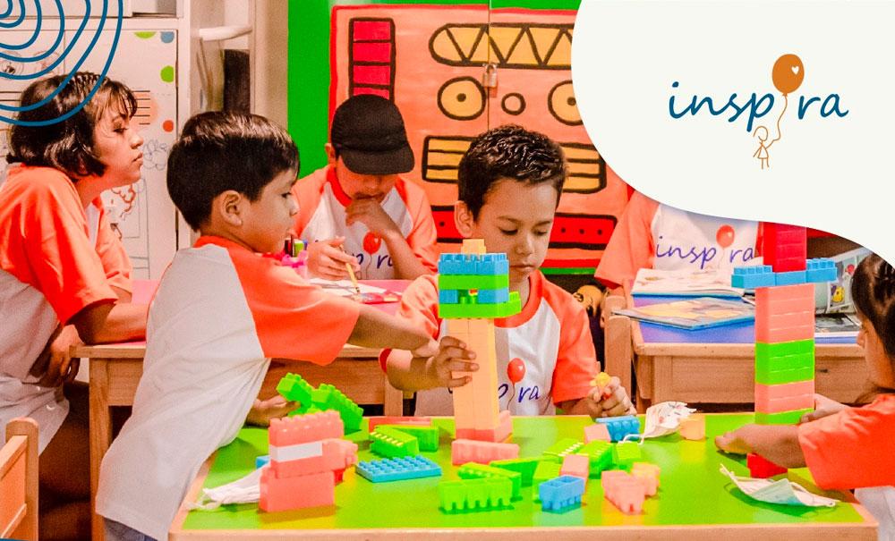 Three children sitting at a green table playing with large legos. There are two other tables behind with other children playing